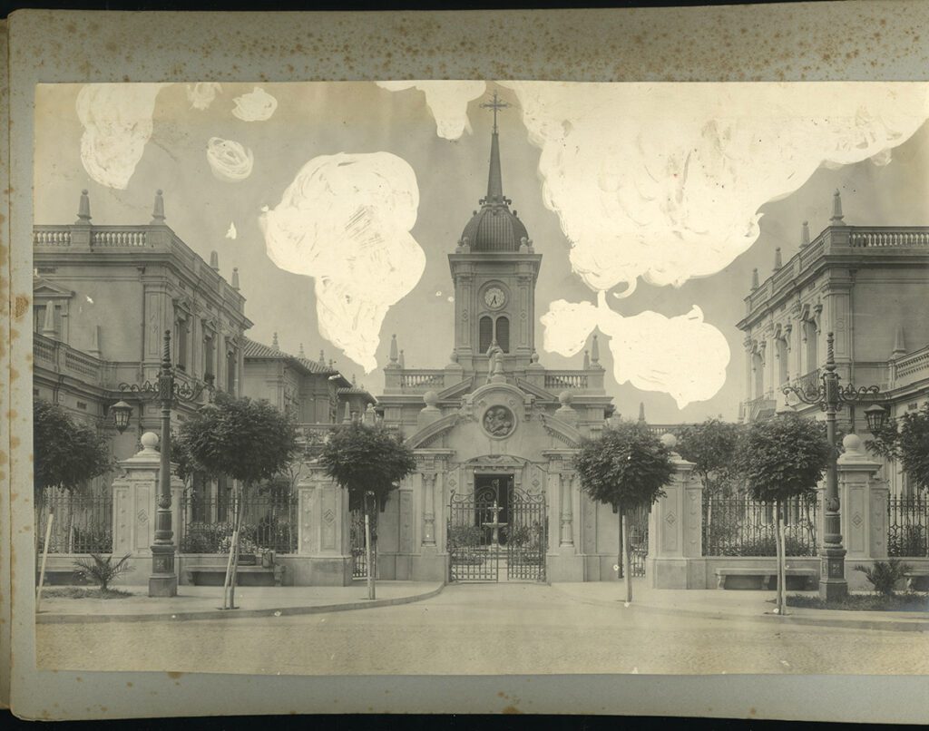 Imagen en blanco y negro de la Iglesia Santa Lucrecia. En el centro se observa la cúpula de la parroquia. A los costados están la Gota de Leche, izquierda, y Casa de Acogida, derecha. En frente de la edificación se encuentra una pileta de mármol. En primer plano, se ve la reja y muralla que rodea la construcción y algunos árboles medianos en la vereda. La fotografía está puesta sobre papel gris de álbum de fotos y tiene algunas manchas blancas.