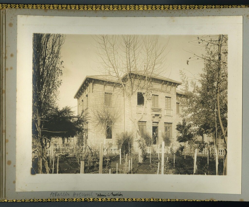 Frontis del edificio de la Caja de Ahorro en blanco y negro. En primer plano se ven árboles y plantas de la Plaza Huemul, tras ellos se ve el edificio con la puerta principal abierta.