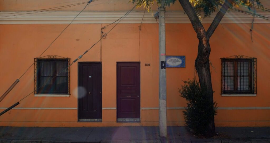 Frontis de casa de hormigón de un piso que sigue el diseño de las casas de la Población Huemul I. La pared es de color damasco y se observa una placa al lado izquierdo de la puerta de calle.