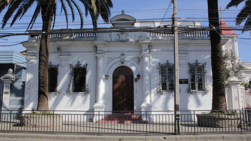 Frontis del edificio actual de la ex Gota de Leche, el cual hoy es una residencia de Mejor Niñez (Autoría: María Francisca Escobar). Frontis actual del edificio de la ex Gota de Leche. Construcción de un piso con una puerta al centro y ventanas a los lados de la fachada. La fachada es color blanco y está flanqueada por dos palmeras.