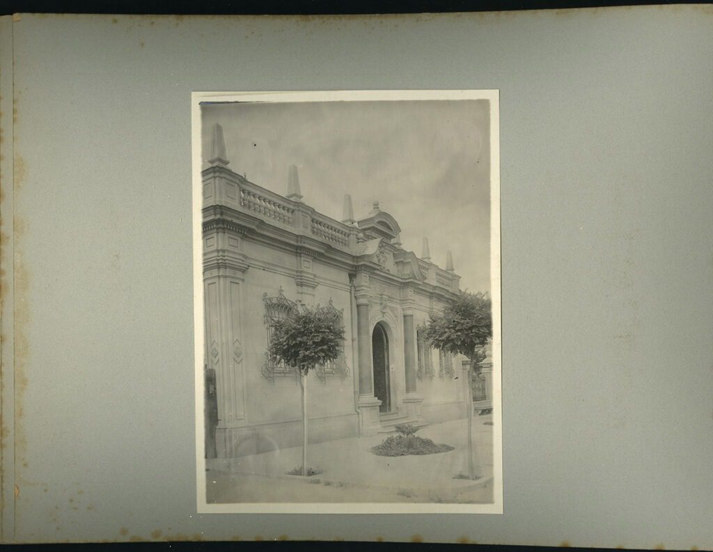 Frontis de la Gota de Leche a principios del siglo XX (Fuente imagen: Población Huemul Inauguración de la sección beneficencia). Imagen en blanco y negro del frontis del edificio de la Gota de Leche. La fotografía está puesta sobre un papel gris de álbum de fotos. El frontis es de color claro, la construcción posee un piso y se ven algunos árboles pequeños en la vereda.