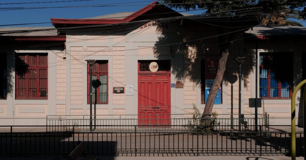Frontis del edificio de la ex Escuela de Mujeres. Se mantiene la construcción de un piso, con paredes de hormigón, ventanas a lo largo del frontis y una puerta principal. La calle es de cemento y se ven árboles en la vereda. Al lado de la puerta principal se observan placas con el nombre del establecimiento.