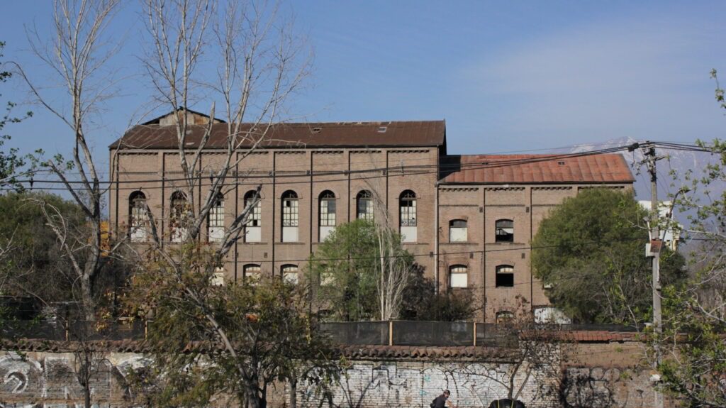 Fotografía del lado poniente del edificio de la ex Refinería de Azúcar. El plano es general y se ve gran parte del edificio el cual posee una muralla alta alrededor, y tras esta un edificio alto con grandes ventanas, algunas están rotas.