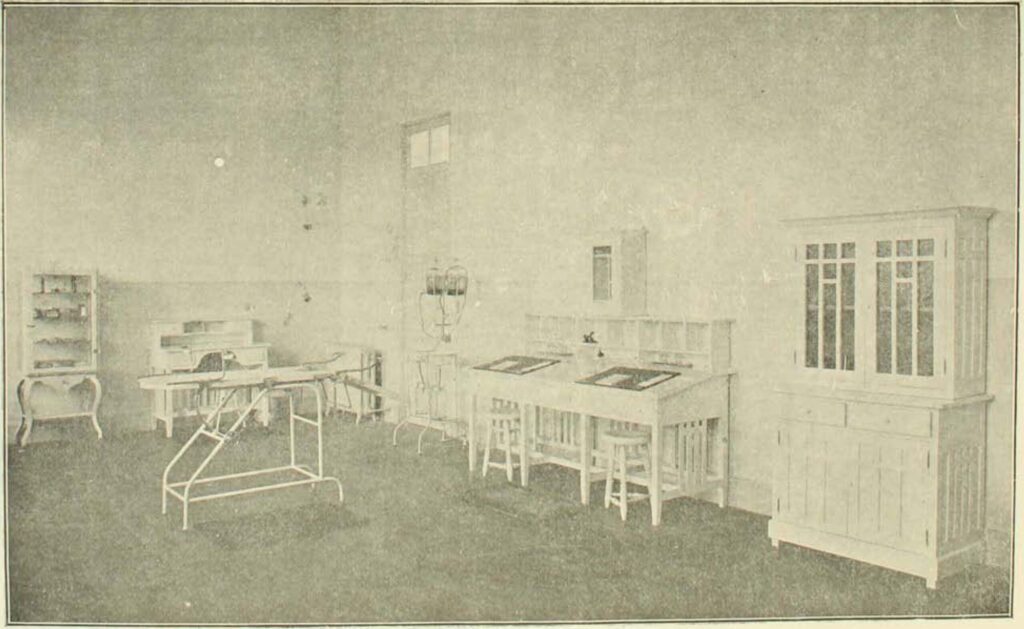 Interior del edificio de la Gota de Leche, específicamente, la sala de curaciones (Fuente imagen: Población Huemul Inauguración de la sección beneficencia, 1918). Imagen en blanco y negro de la sala de curaciones del edificio de la Gota de Leche. Se observa una sala amplia, con implementos tales como camilla, vidrieras, mesones de trabajo y otros. Murallas y techos altos con colores claros.