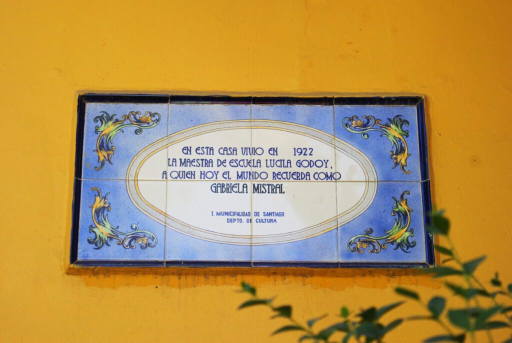 Imagen en colores de placa de cerámica ubicada en el frontis de la casa de Gabriela Mistral en la actualidad. La pared es amarilla y en el centro de la imagen se observa la plaza que dice: En esta casa vivió en 1922 la maestra de escuela Lucila Godoy, a quien hoy el mundo recuerda como Gabriela Mistral. Ilustre Municipalidad de Santiago, Departamento de Cultura.