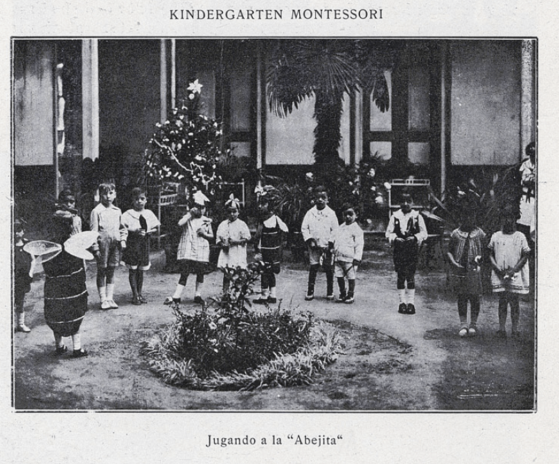 Grupo de Kindergarten Montessori jugando a la “Abejita”. Año 1927. Fotografía en blanco y negro de niños y niñas que juegan en círculo en un patio (Copyright: Patrimonio Cultural Común, extraída del libro Actividades Femeninas en Chile, disponible en http://www.memoriachilena.gob.cl/602/w3-article-126684.html).