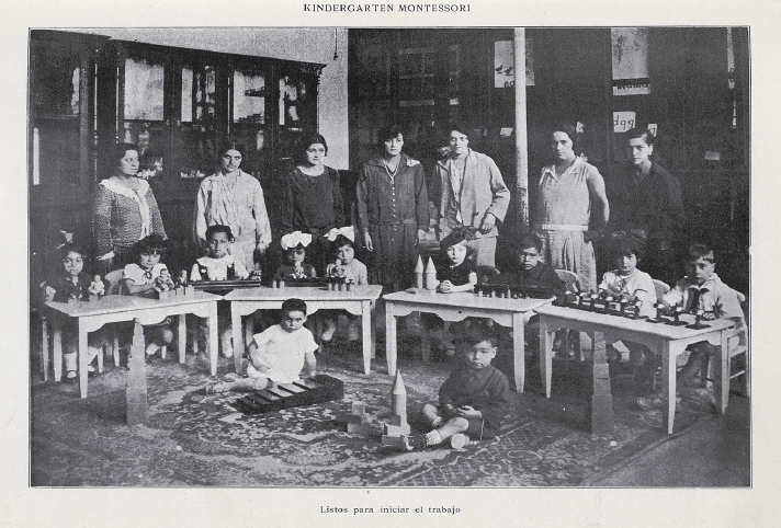 Grupo de niños, niñas y educadoras de Kindergarten Montessori. Año 1927. Fotografía en blanco y negro, niños y niñas estñan sentados en el suelo o en sillas y tienen juguetes en las mesas. Las educadoras están de pie detrás de los niños y niñas (Copyright: Patrimonio Cultural Común, extraída del libro Actividades Femeninas en Chile, disponible en http://www.memoriachilena.gob.cl/602/w3-article-126684.html).